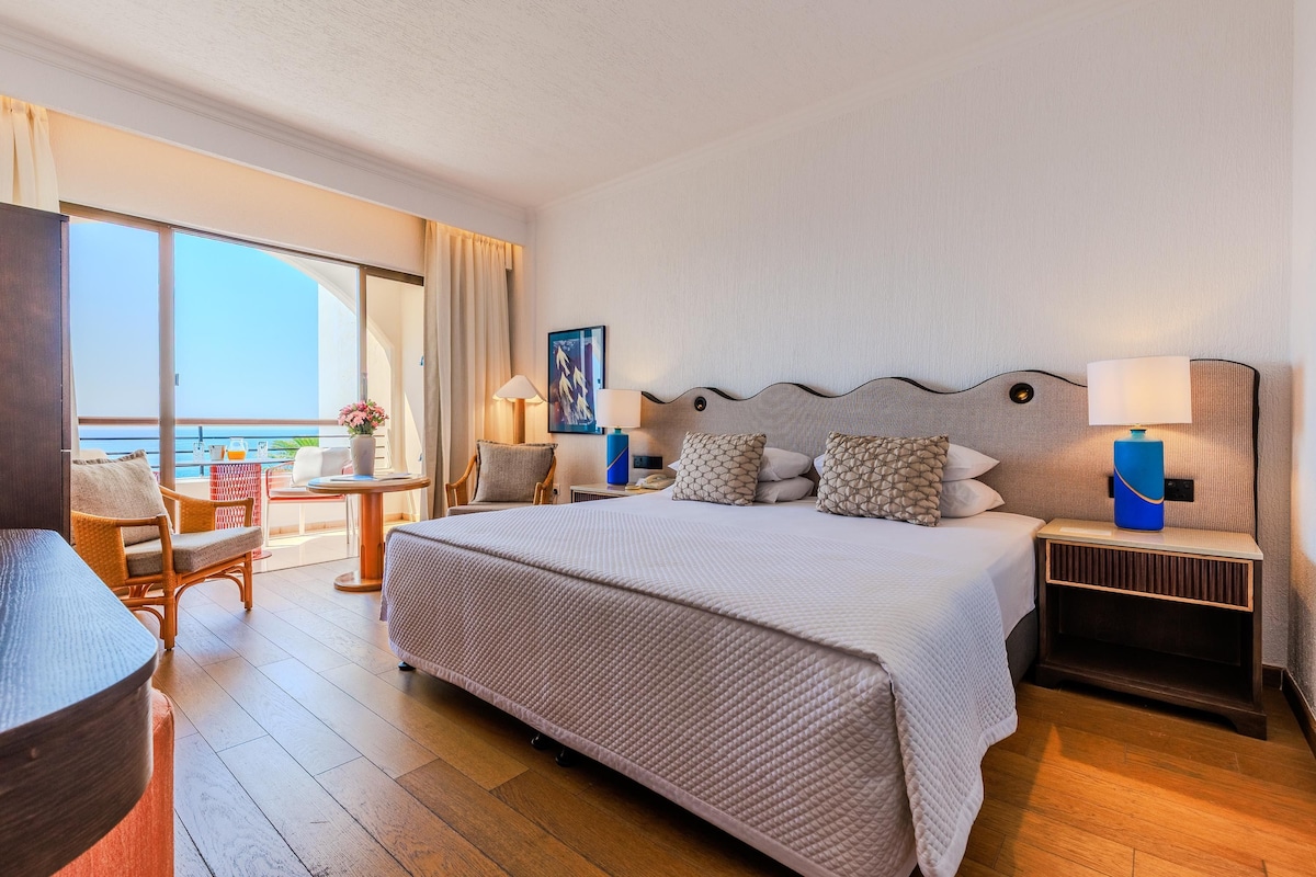 Quarto de hotel com uma cama grande, uma escrivaninha, uma cadeira e vista para o oceano.