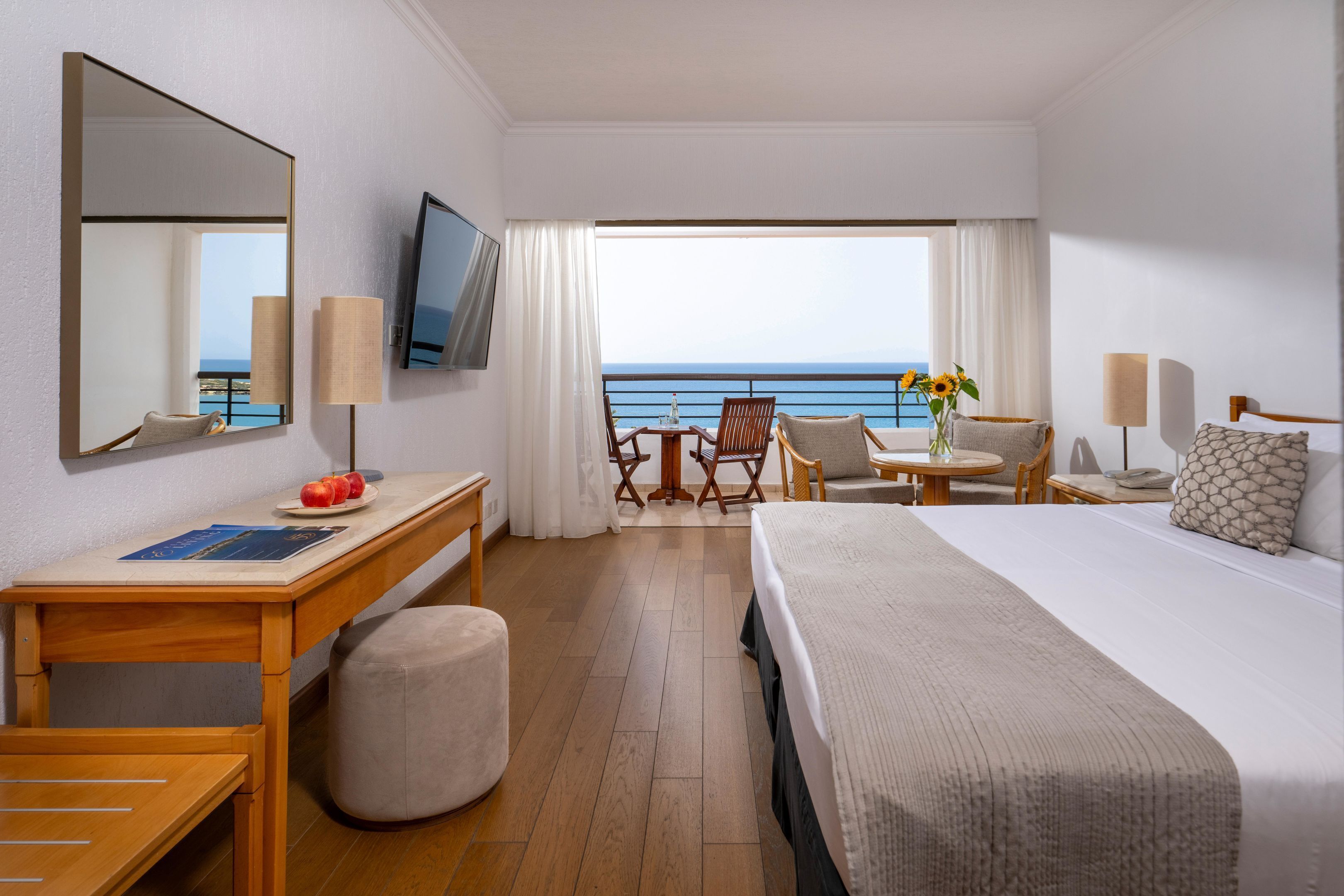A hotel room with a bed, desk, chairs, a TV, and a view of the ocean.