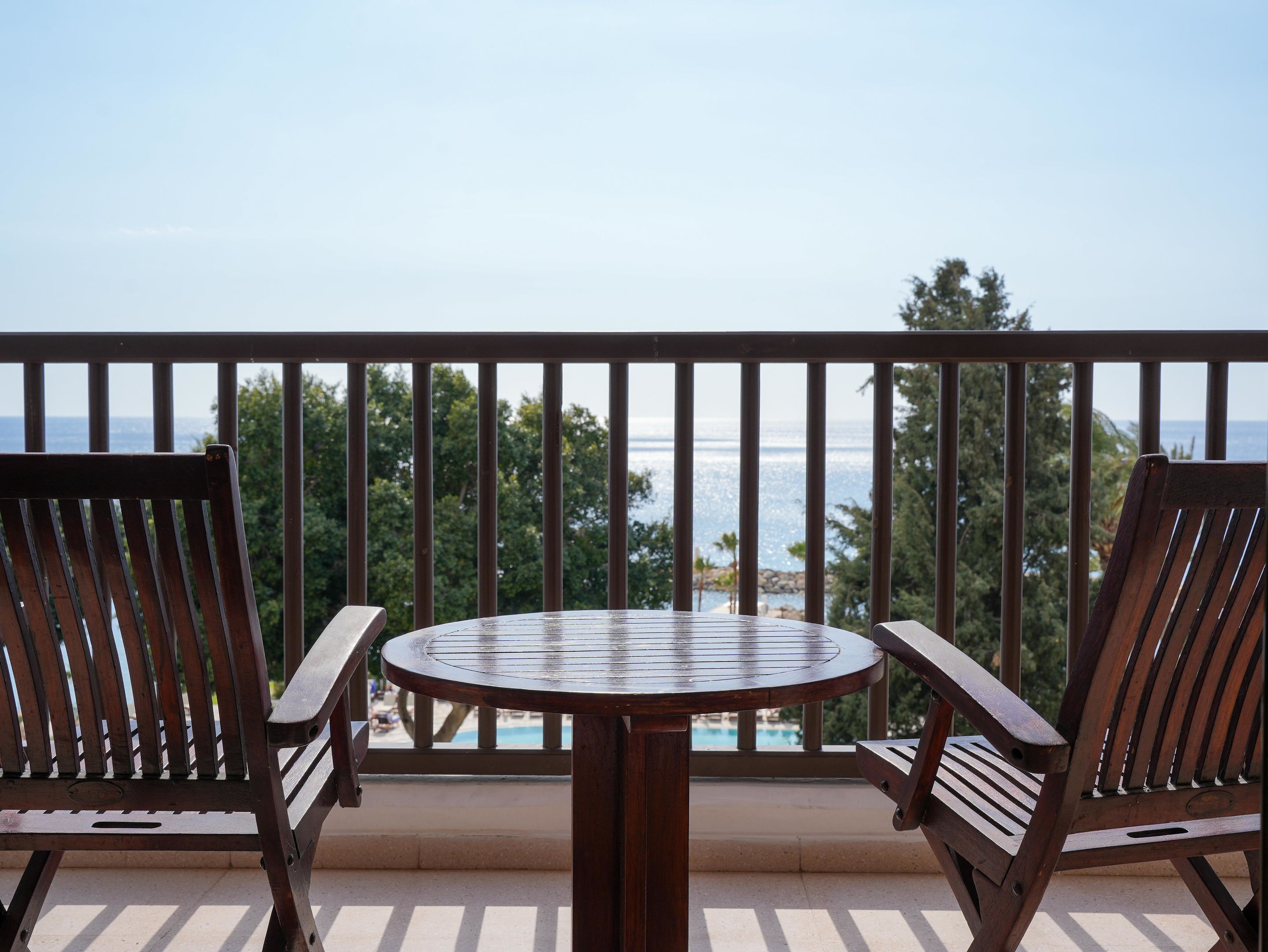 A balcony with a round wooden table and two wooden chairs, overlooking a body of water and greenery.