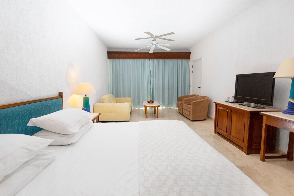 Een hotelkamer met een bed, een bureau met een televisie, een stoel, een bijzettafeltje met een lamp, en een plafondventilator.