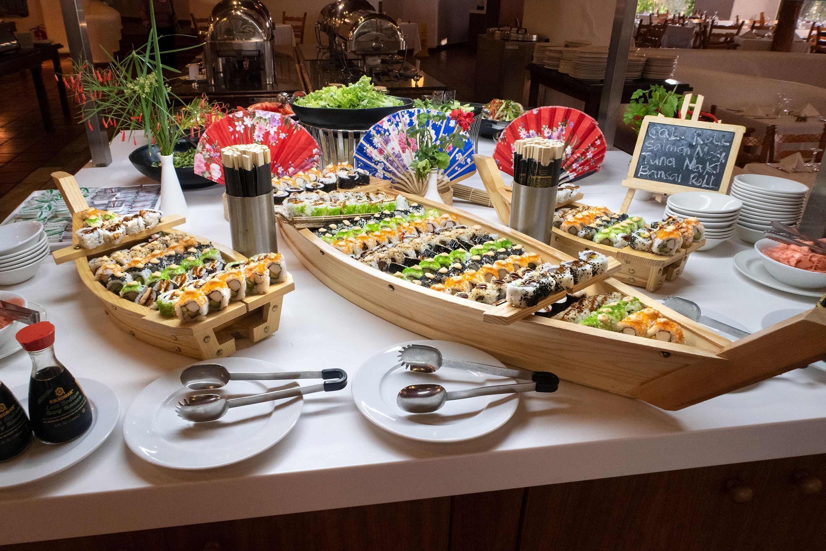 A buffet table with various sushi dishes, chopsticks, plates, and a sign indicating available rolls.