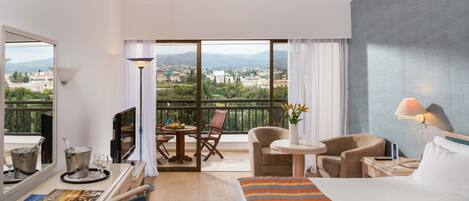 Habitación de hotel con una cama grande, un escritorio con computadora, una silla, una mesita con lámpara y un balcón con vistas a la ciudad.