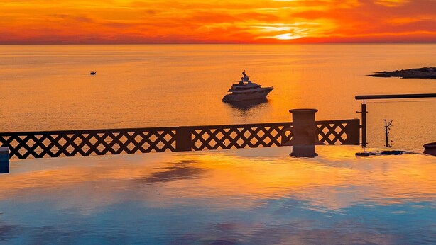 A sunset over a calm sea with a yacht in the distance, a wooden deck, and a railing. - Coral Beach Hotel and Resort (Pegeia)