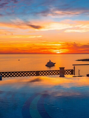 A sunset over a calm sea with a yacht in the distance, a wooden deck, and a railing. - Coral Beach Hotel and Resort (Pegeia)