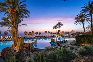 Zona de piscina con sillas de descanso, palmeras y vista al océano al atardecer.
