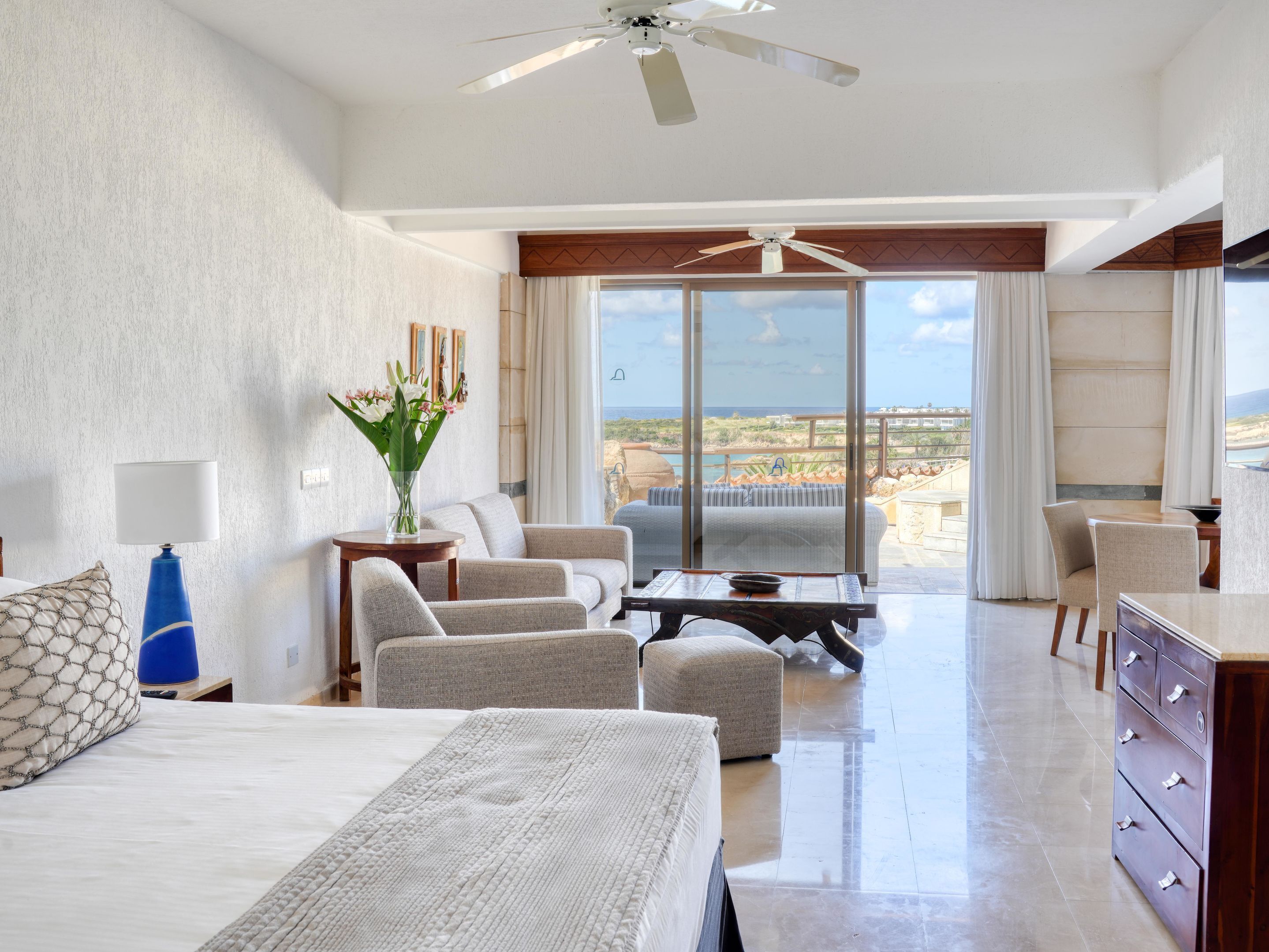 Ein Hotelzimmer mit Bett, einer Sitzecke mit Stühlen, einem Tisch, einer Vase mit Blumen und Blick aufs Meer.