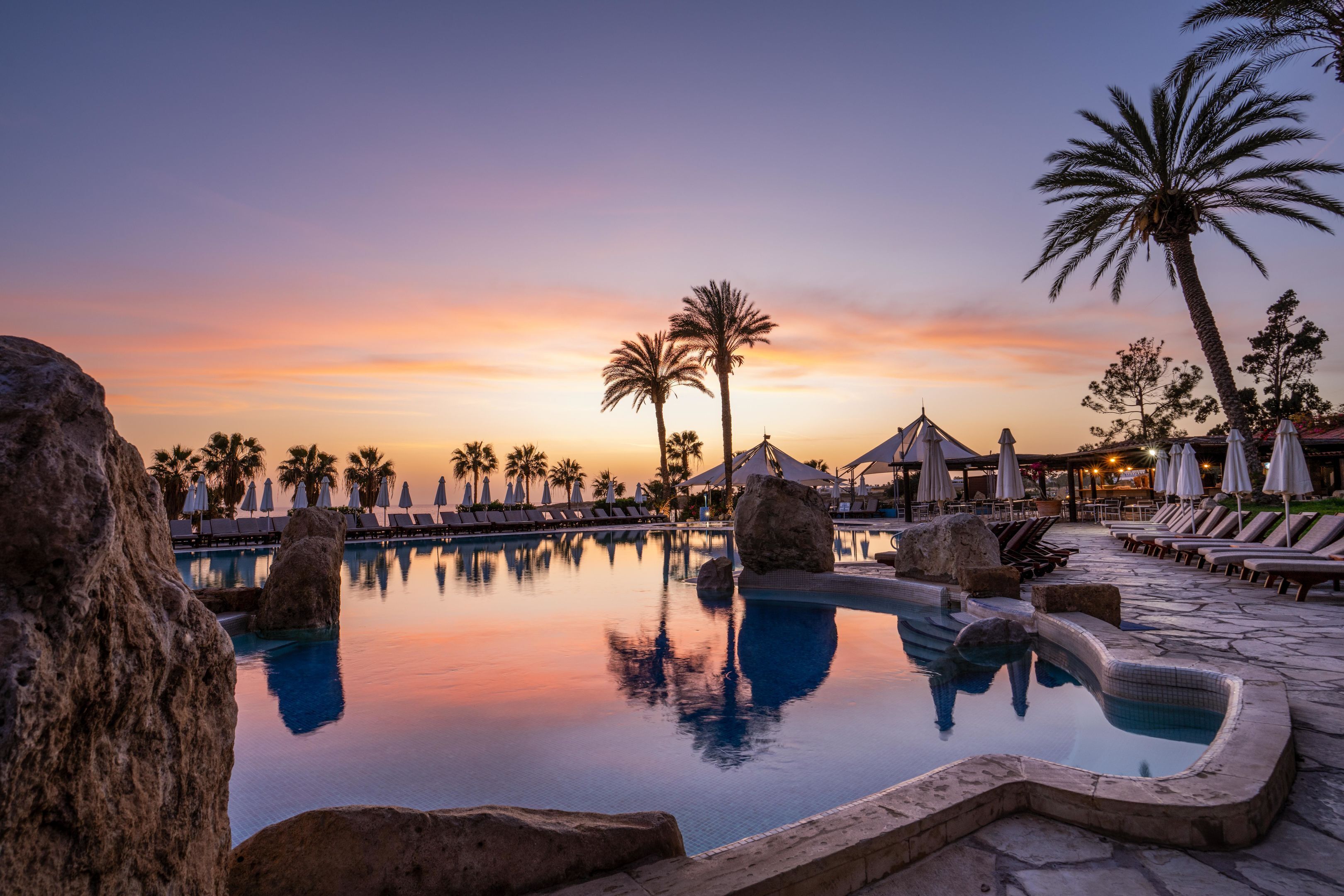Zona de piscina con sillas de descanso, palmeras y un cielo al atardecer.