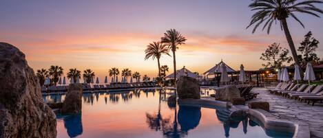 Zona de piscina con sillas de descanso, palmeras y un cielo al atardecer.