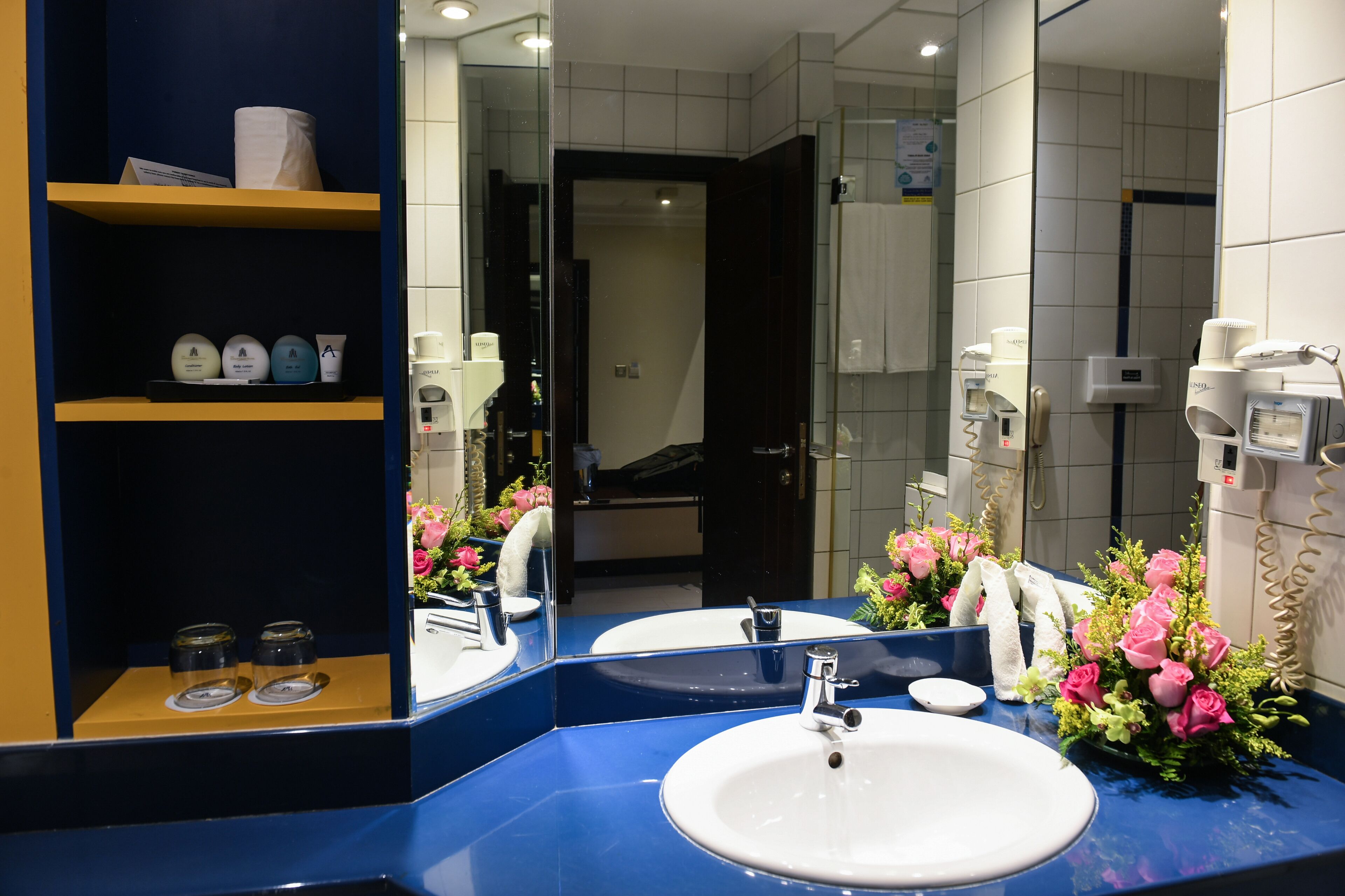 A bathroom with a blue vanity, white sink, and a mirror reflecting the room. There are shelves with toiletries and a shower area visible in the background.
