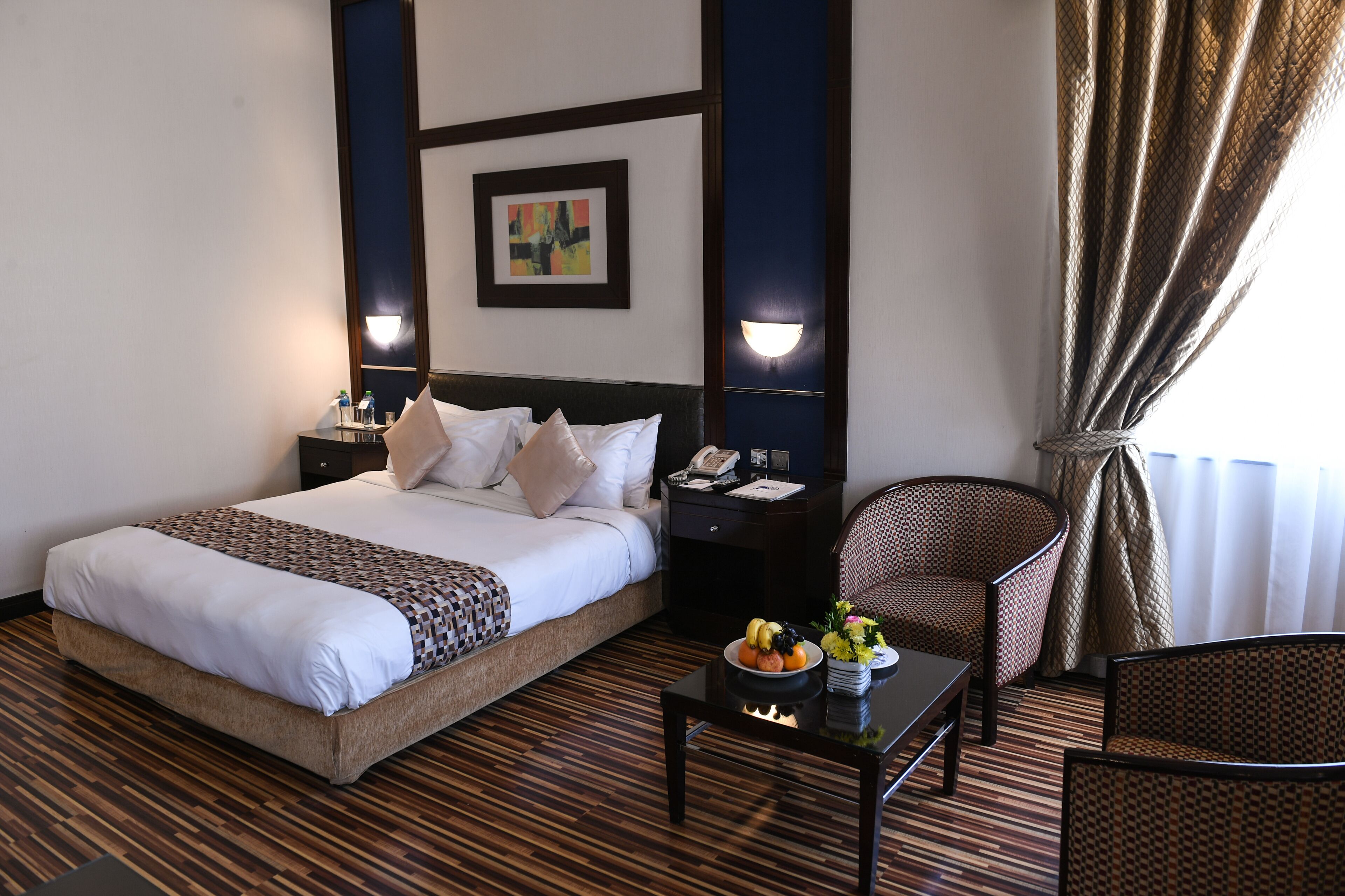 A hotel room with a large bed, a bedside table with a phone, a chair, a small table with fruit, and a wall-mounted light fixture.