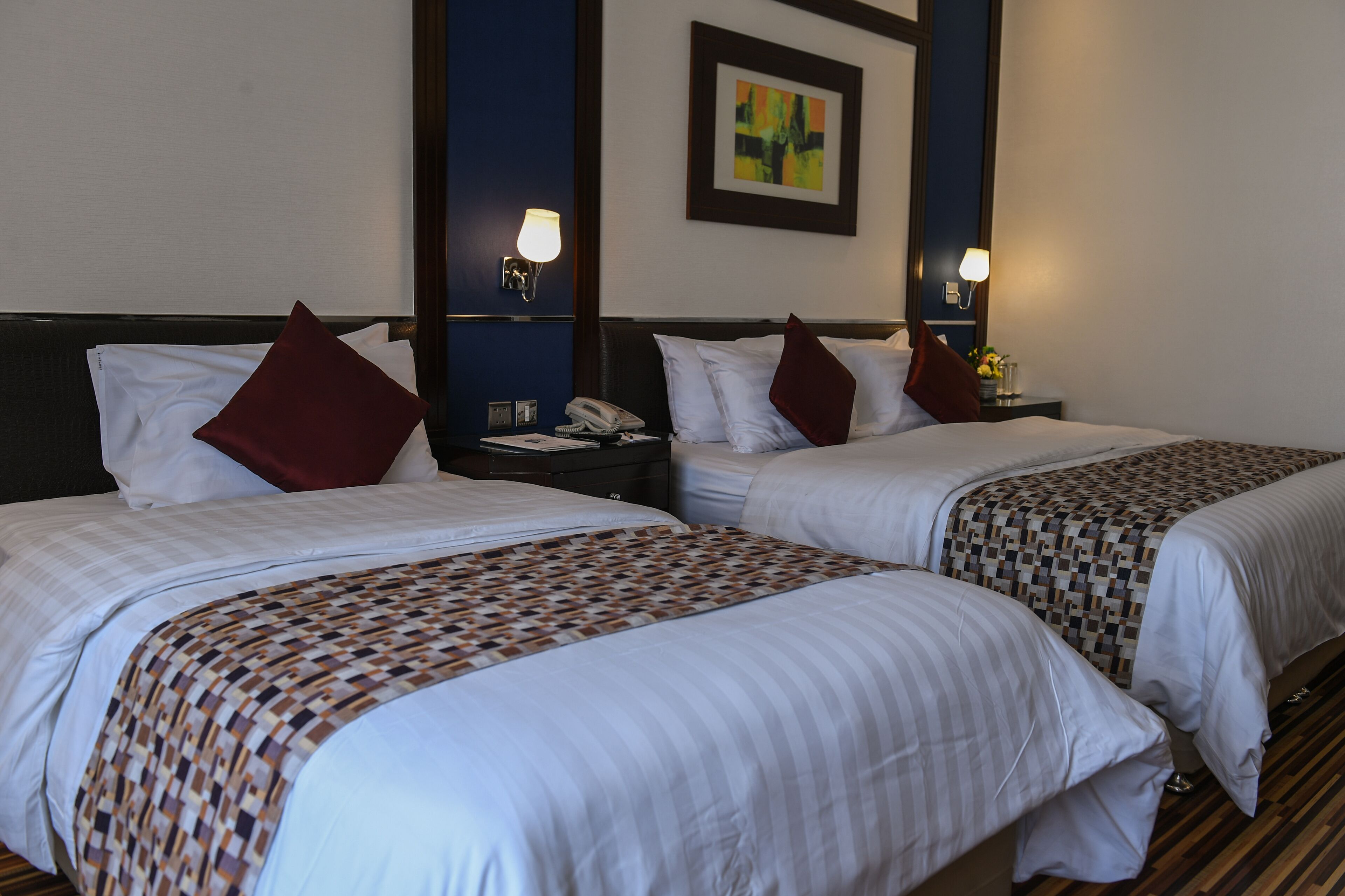 Habitación de hotel con dos camas, cada una con cubrecama estampado, ropa de cama blanca y almohadas rojas. Hay lámparas de noche, un teléfono en la mesita de noche y una obra de arte enmarcada en la pared.
