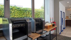 A kitchen area with commercial ovens, a buffet table with food items, and a view of a green hedge outside. - Holiday Inn Express London - Dartford by IHG (Dartford)