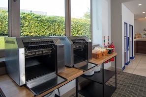 A kitchen area with commercial ovens, a buffet table with food items, and a view of a green hedge outside. - Holiday Inn Express London - Dartford by IHG (Dartford)