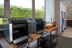 A kitchen area with commercial ovens, a buffet table with food items, and a view of a green hedge outside.