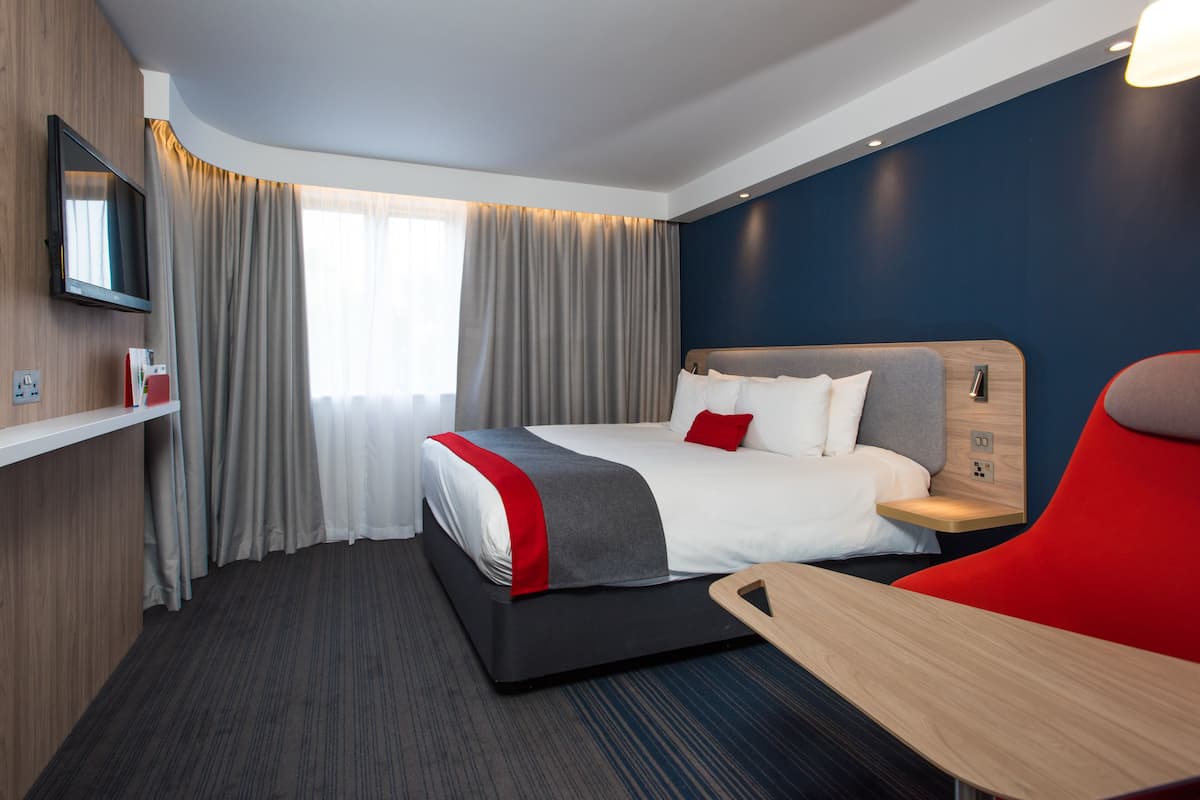 Una habitación de hotel moderna con una cama grande, una mesita de noche, un televisor en la pared y una ventana con cortinas.