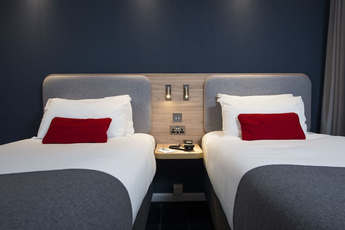 A hotel room with two beds, each with a grey headboard, white bedding, and a red pillow. There is a wooden panel between the beds with a small table and a phone.