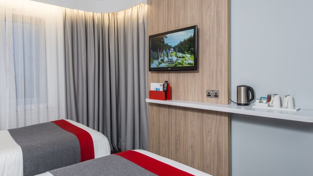 A hotel room with two beds, a TV mounted on the wall, a wooden panel, a shelf with a kettle and cups, and a curtain. - Holiday Inn Express London - Dartford by IHG (Dartford)