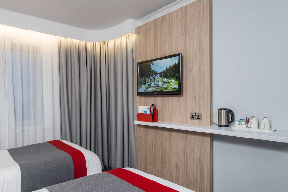 Habitación de hotel con dos camas, un televisor montado en la pared, un panel de madera, un estante blanco con un hervidor y tazas, y una colcha de color rojo y gris.