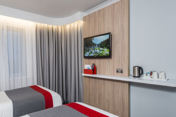 A hotel room with two beds, a TV mounted on the wall, a wooden panel, a shelf with a kettle and cups, and a curtain. - Holiday Inn Express London - Dartford by IHG (Dartford)