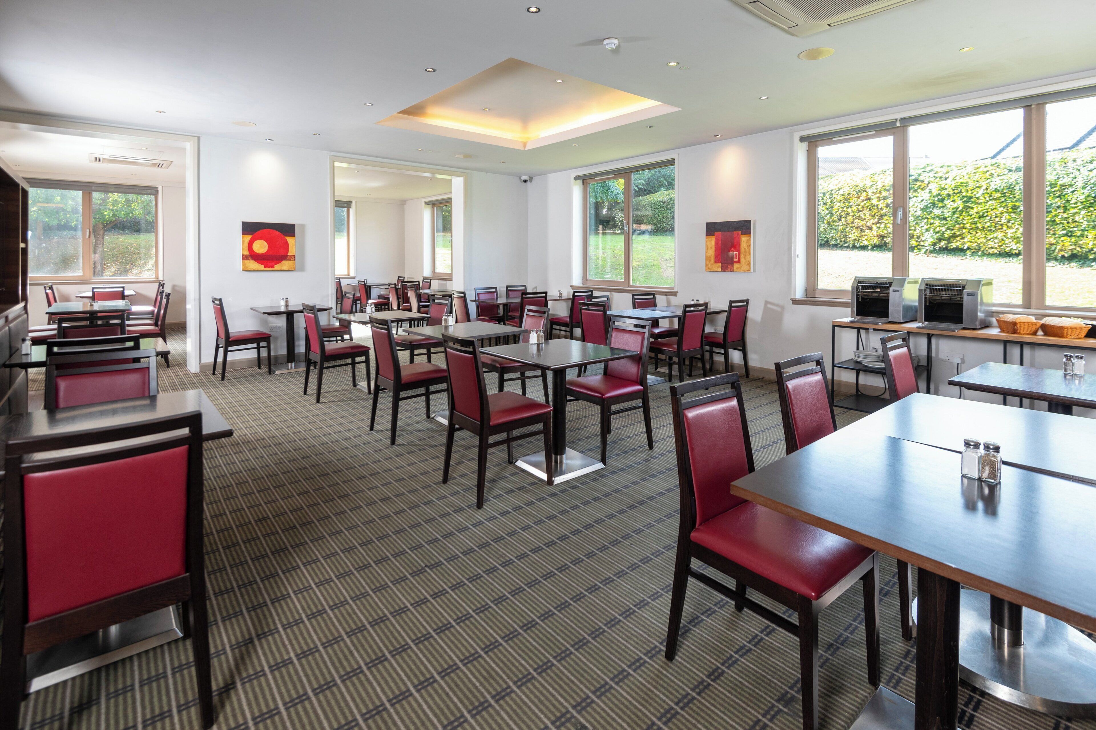 Une salle à manger d’hôtel avec de nombreuses tables et chaises, de grandes fenêtres et une vue sur la verdure environnante.
