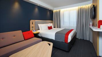 A hotel room with a bed, a sofa, a small table, a TV mounted on the wall, and a window with curtains.