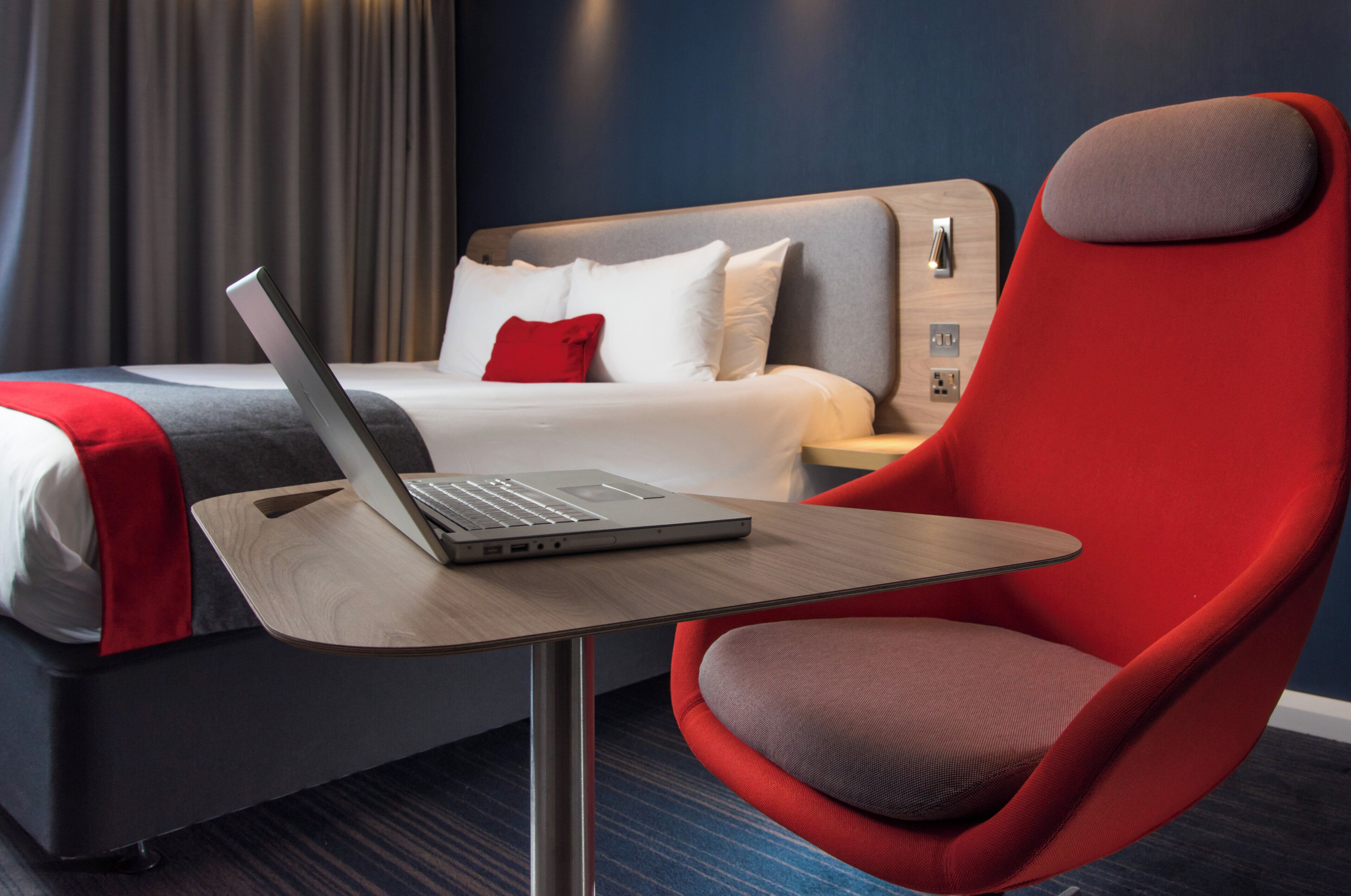 Une chambre d’hôtel avec un lit, un fauteuil rouge, une table en bois avec un ordinateur portable et une table de chevet avec une lampe.