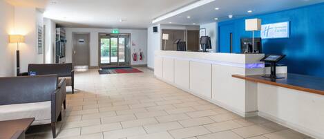 A hotel reception area with a blue wall, a reception desk, a seating area with a sofa and chairs, and a tiled floor.
