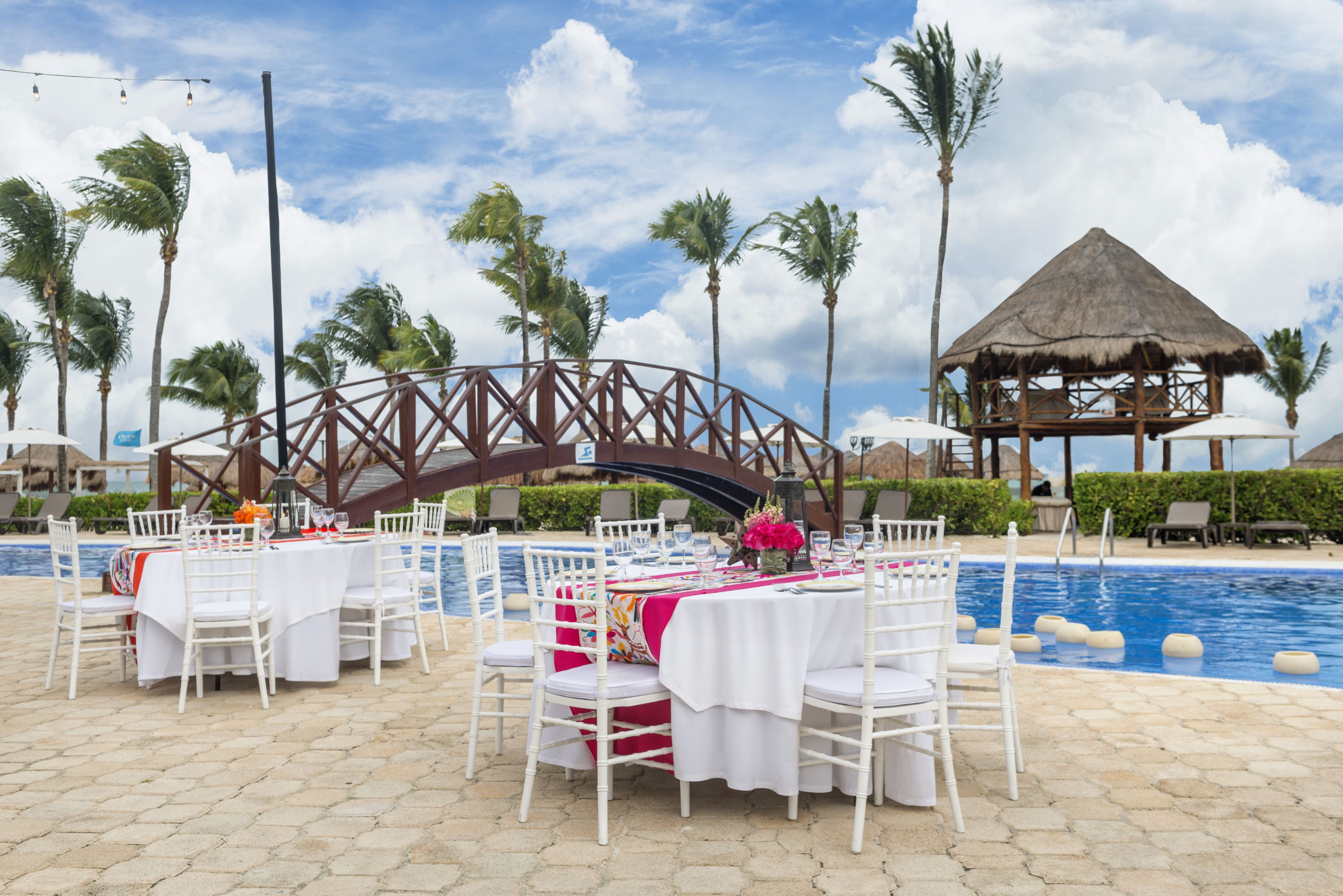 A poolside dining area with white tables and chairs, a thatched-roof structure, palm trees, and a bridge in the background.
