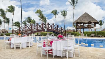 A poolside dining area with white tables and chairs, a thatched-roof structure, palm trees, and a bridge in the background.