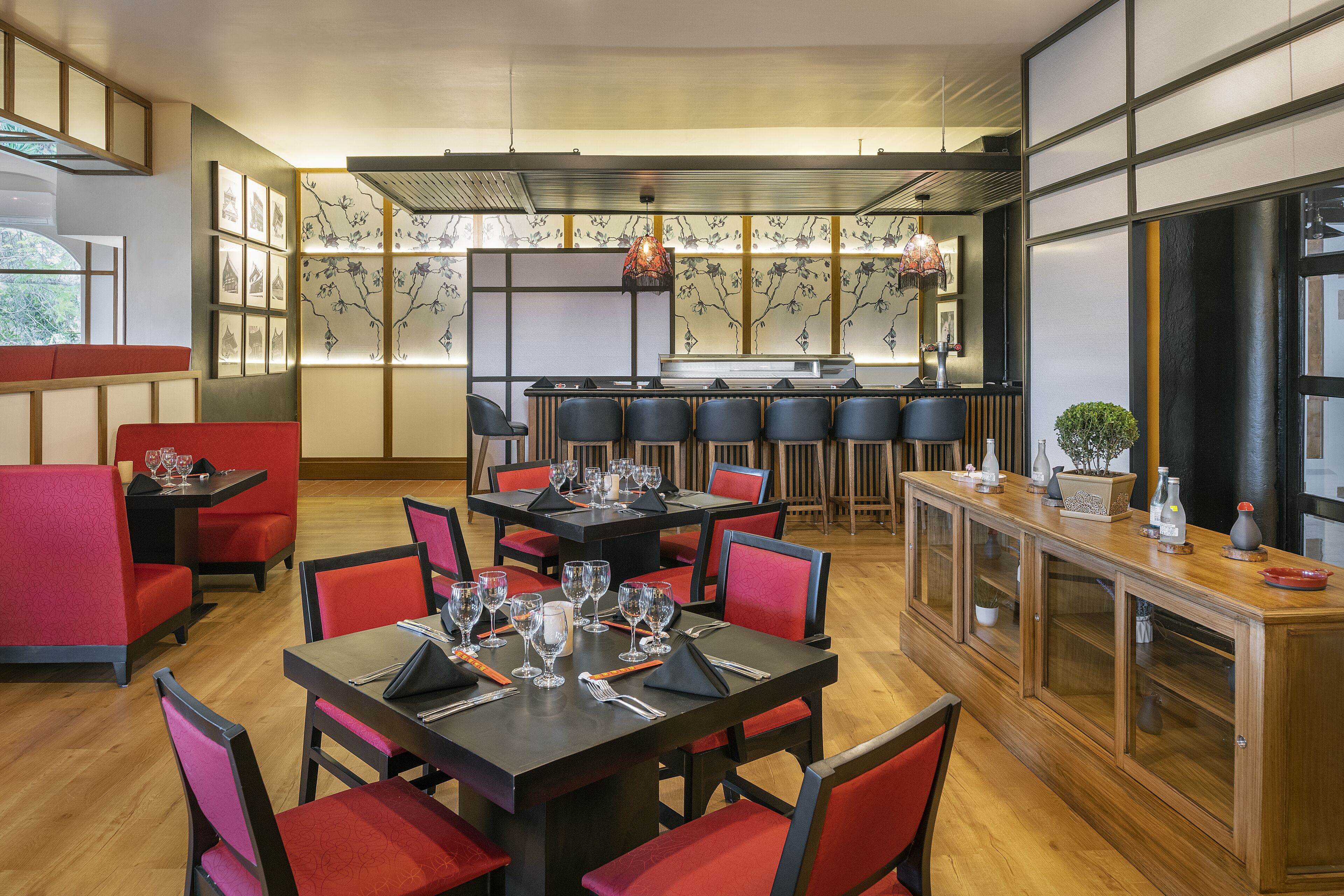 A restaurant with wooden flooring, red and black chairs, and a long counter with a plant and decorative items.
