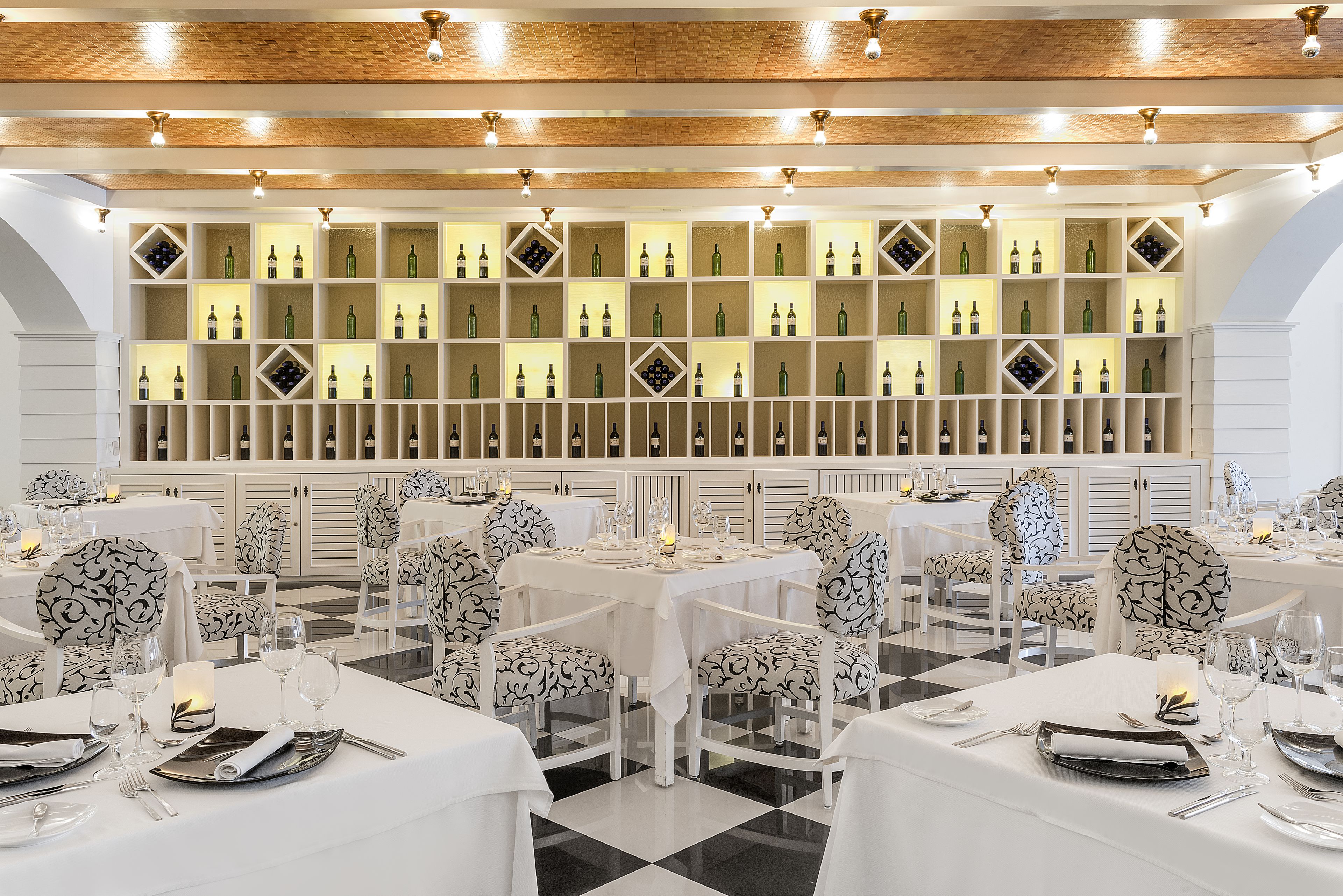 Een restaurant met witte tafels en stoelen, een wand met wijnflessen en een geruit vloertapijt.