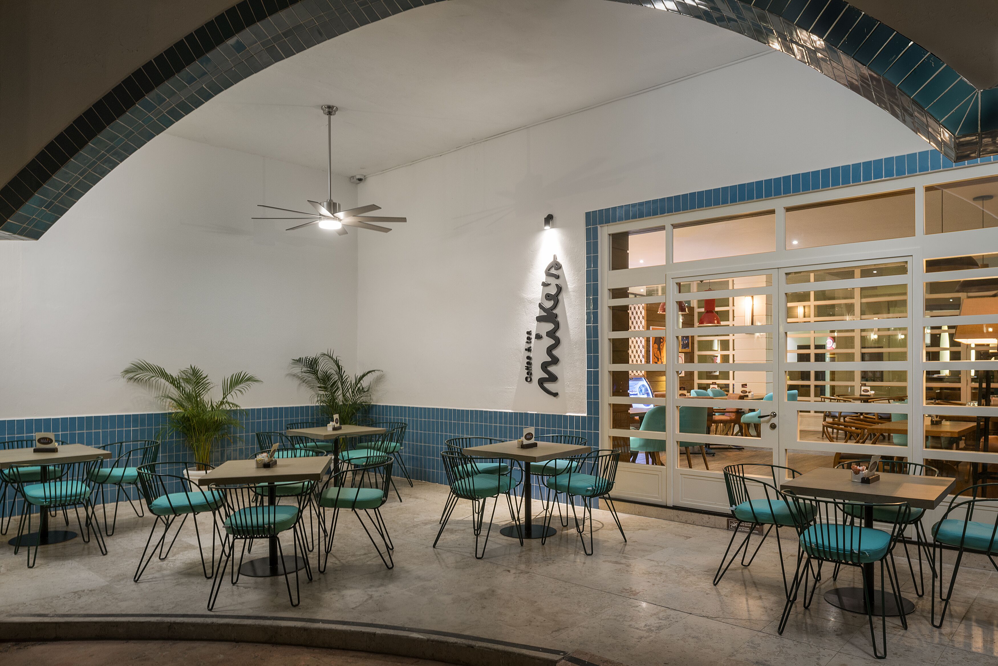 Een restaurant met smaragdgroene stoelen en tafels, een plafondventilator en een groot raam met uitzicht op het interieur.