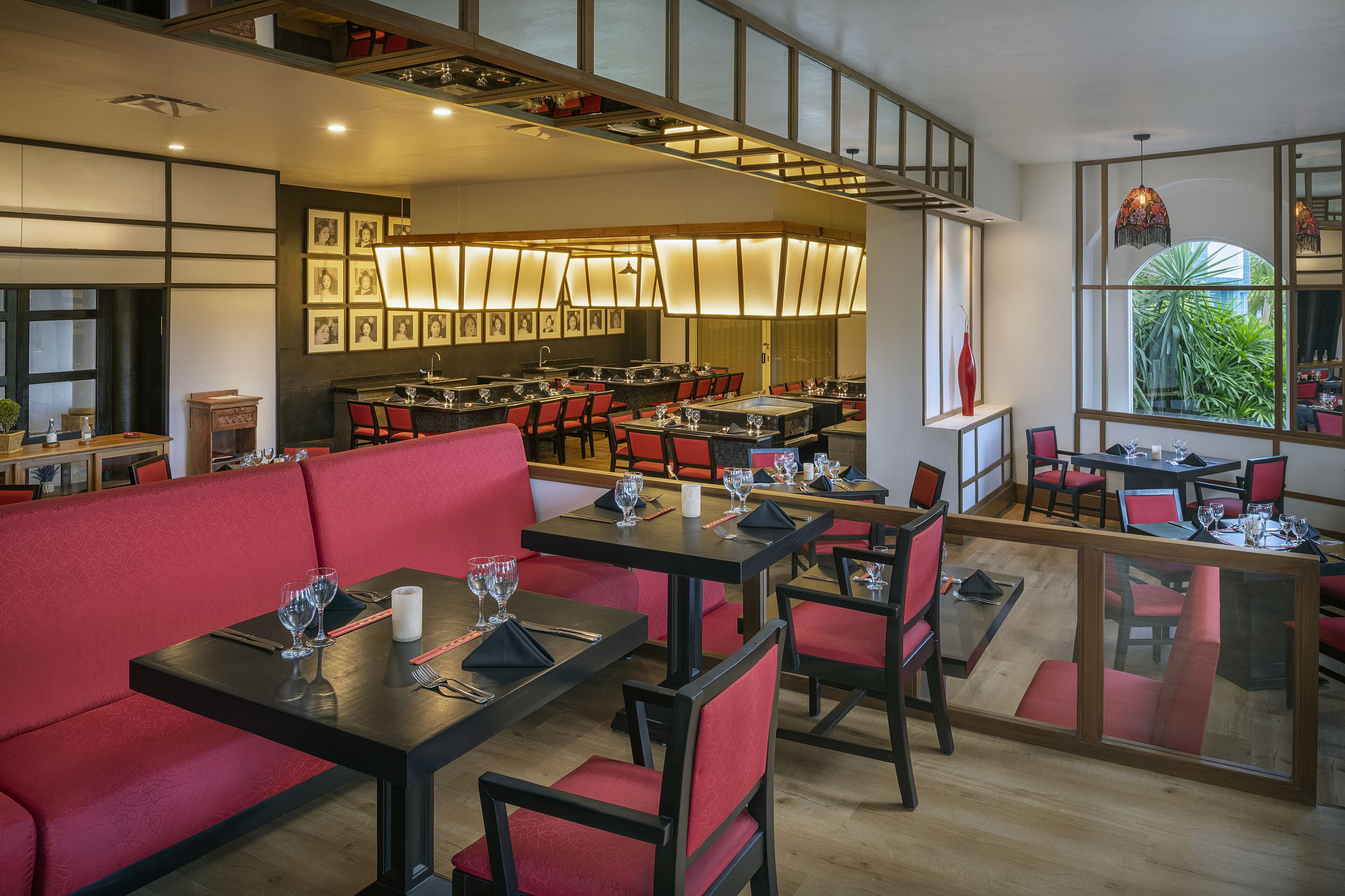 Een restaurant met rode zitplaatsen, meerdere eettafels en uitzicht op groen door grote ramen.