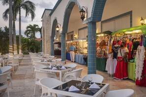 Guests can dine al fresco under the sheltered arches, surrounded by palm trees and a vibrant market stall.