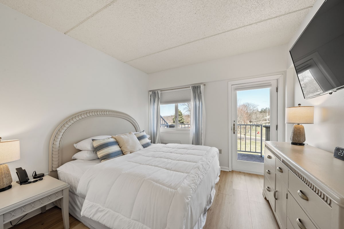 A bedroom with a bed, bedside table, dresser, TV, and a door leading to an outdoor area.
