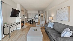 Una sala de estar moderna con un sofá gris, una mesa de centro blanca y una chimenea. Se ve una cocina al fondo con armarios blancos y un comedor.