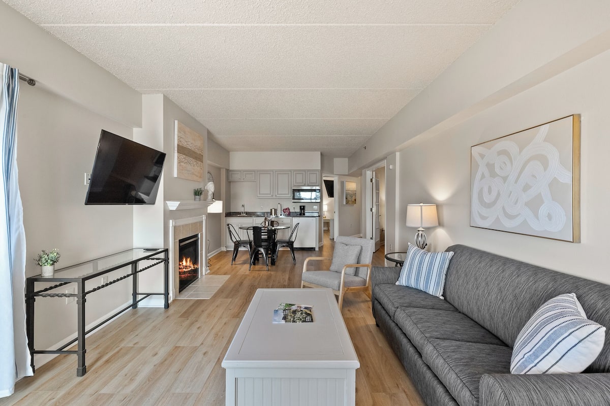 A modern living room with a grey sofa, a white coffee table, and a fireplace. There is a kitchen in the background with white cabinets and a dining area.