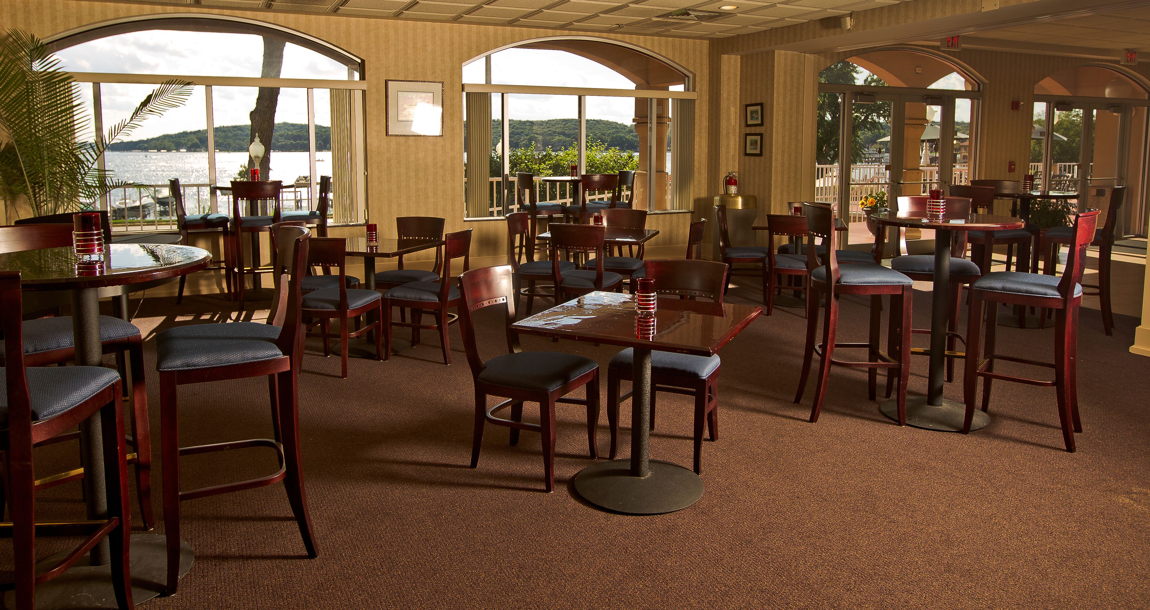 A restaurant with multiple tables and chairs, large windows with a view of the water, and a carpeted floor.