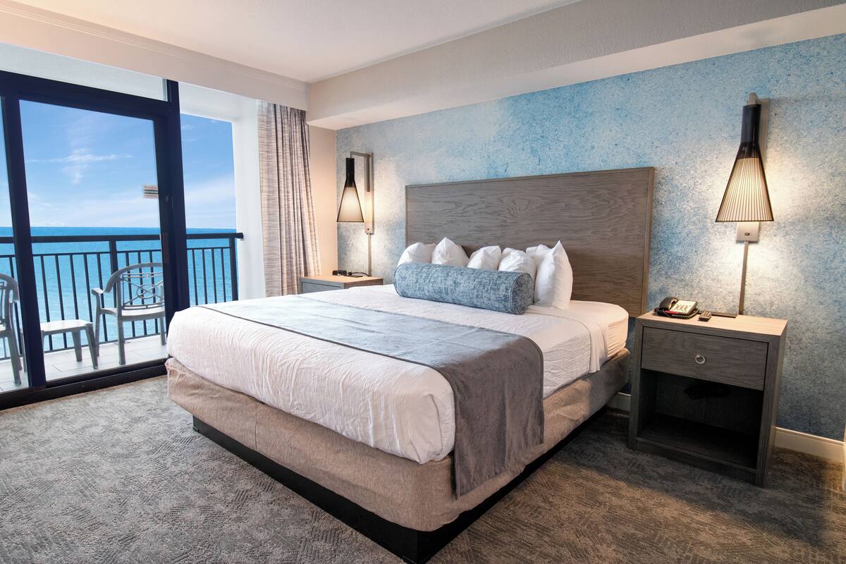 Un'ampia camera da letto con un letto grande, un comodino, una lampada e vista sull'oceano da un balcone.