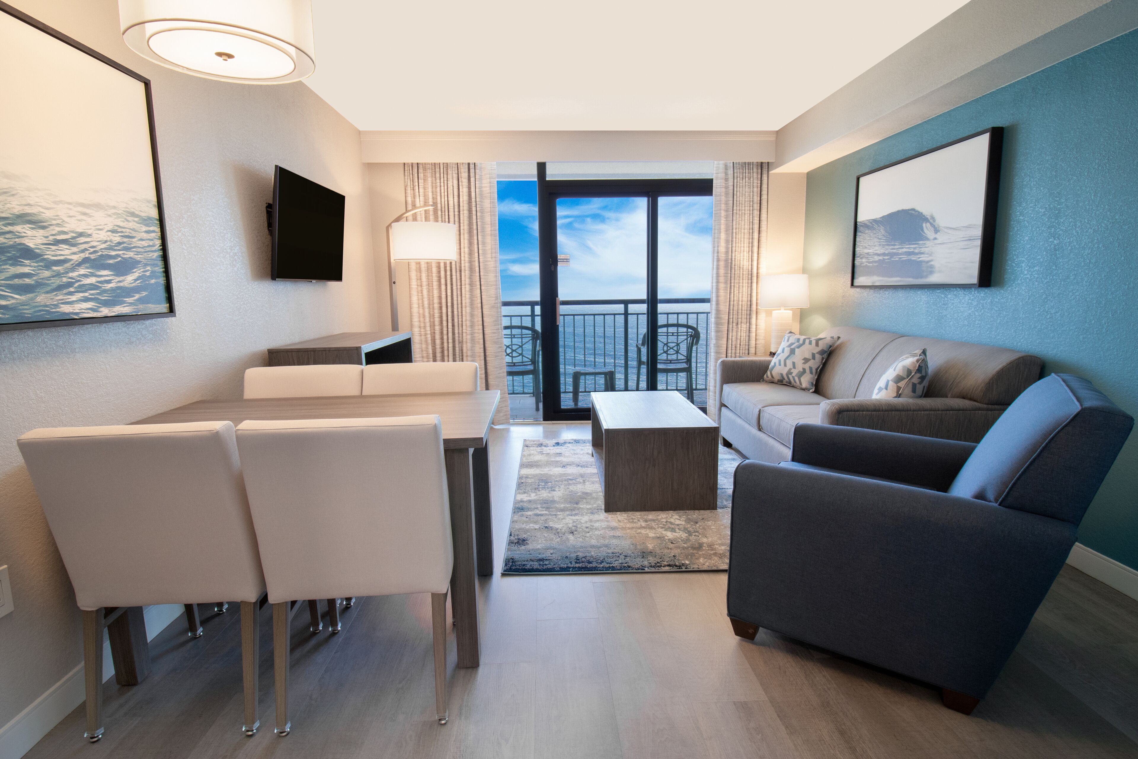 A modern hotel room with a dining area, a sofa, a TV, and a balcony with a view of the sea.