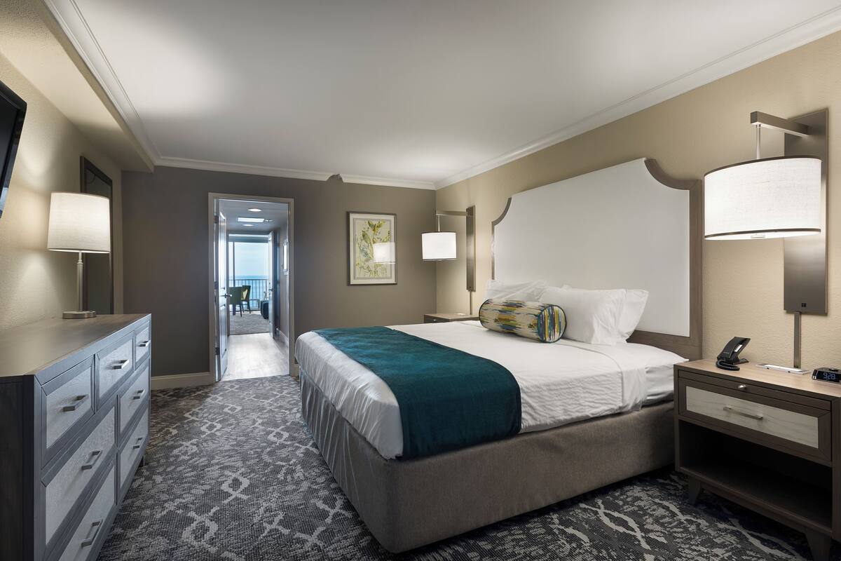 Camera d'albergo con un letto grande, un comodino con una lampada, una TV a schermo piatto fissata al muro e vista sul corridoio e all'esterno.