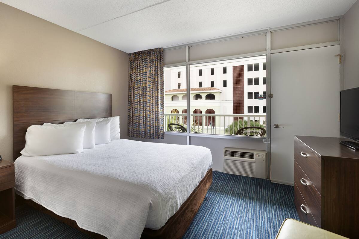Una camera d'albergo con un letto, una scrivania, una sedia, una TV e una finestra che offre vista sugli edifici.
