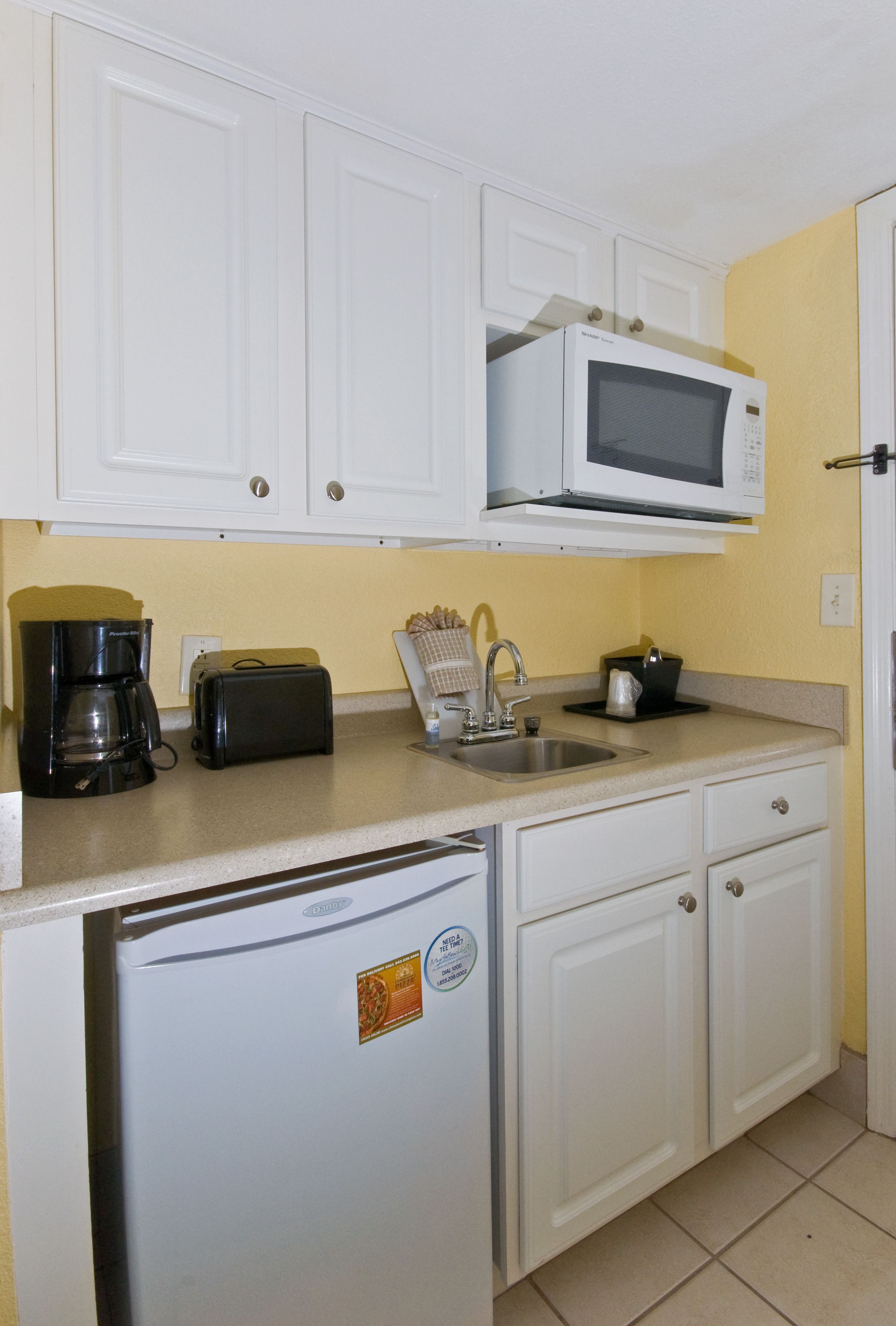 Una cocina compacta con gabinetes blancos, microondas, tostadora, cafetera y refrigerador.
