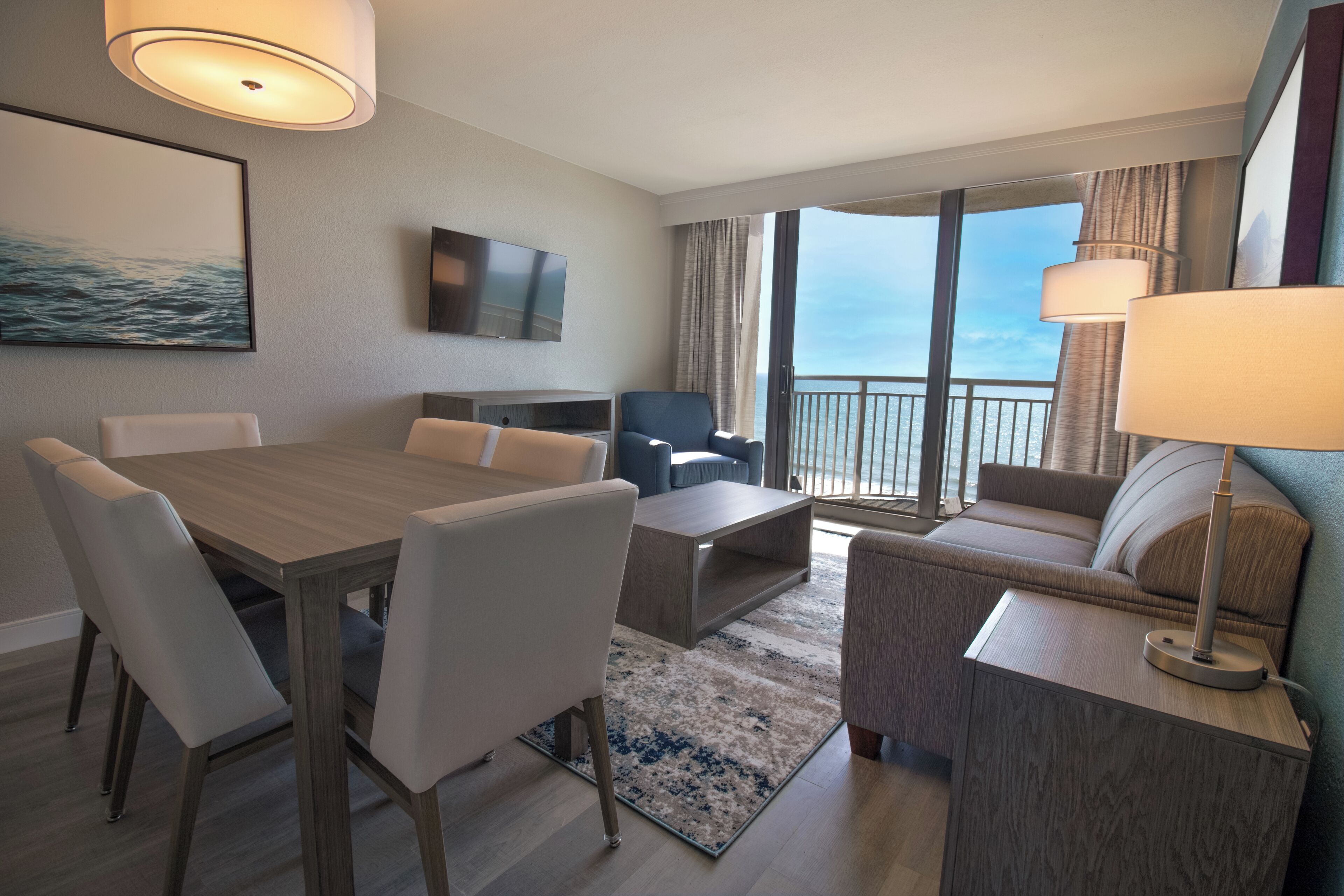 A hotel room with a dining table, chairs, a sofa, a TV, and a balcony with a view.