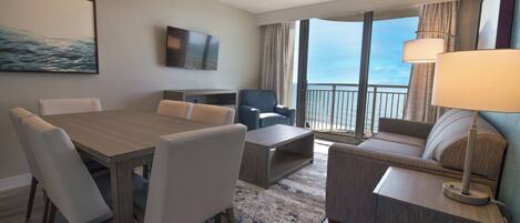 A hotel room with a dining table, chairs, a sofa, a TV, and a balcony with a view.