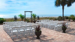 A beachside outdoor seating area with white chairs arranged in rows, a wooden pergola, and potted plants.