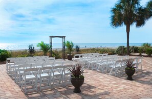 A beachside outdoor seating area with white chairs arranged in rows, a wooden pergola, and potted plants.