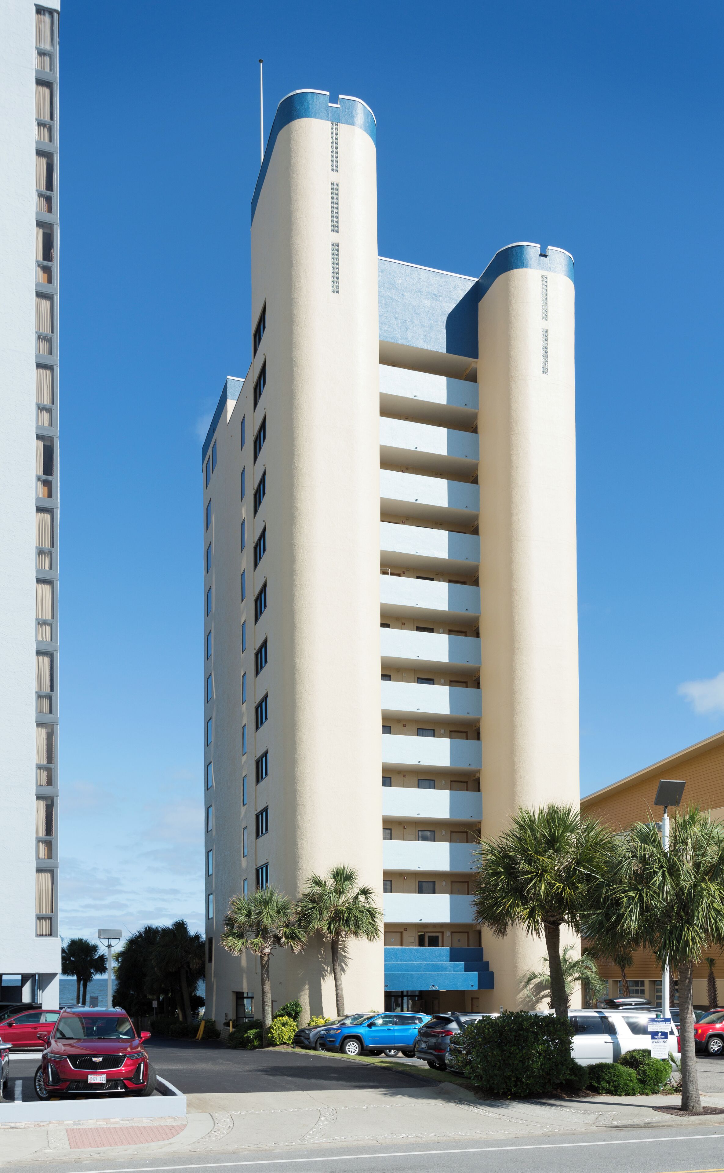 A multi-story hotel with a central cylindrical tower, a parking lot with several cars, and palm trees.