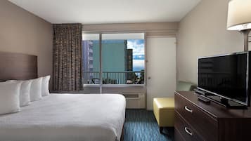 Une chambre d’hôtel comprenant un lit, une télévision, une commode, une fenêtre donnant sur des immeubles et un balcon.