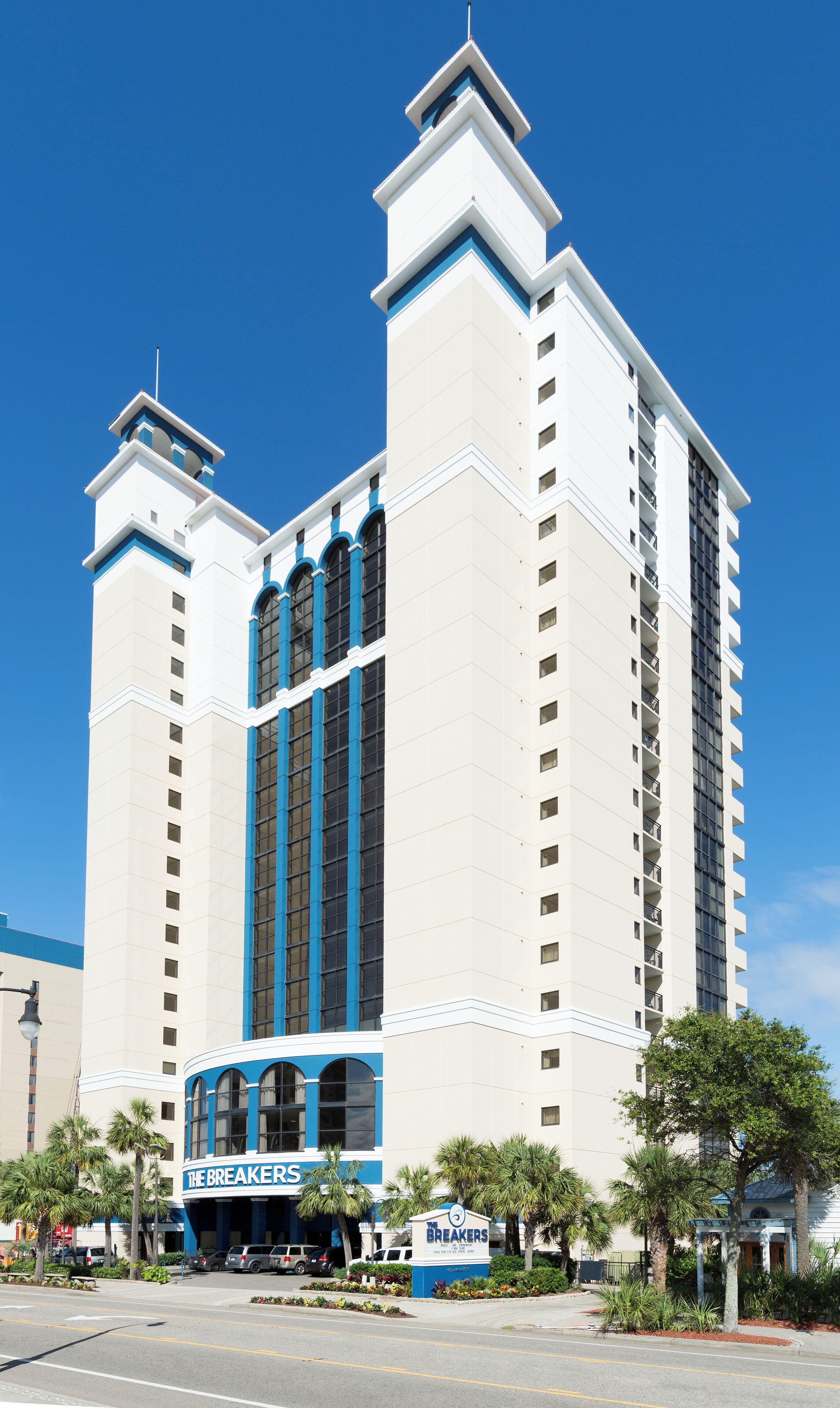A multi-story hotel with a central tower, a sign for 'The Breakers,' and a parking lot with vehicles.